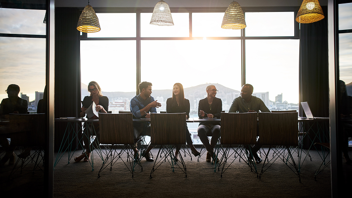 Team in discussion around a long table in a glass-walled office, city skyline and evening light in the background